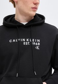 Νεαρό άτομο που φοράει μαύρη φούτερ με κουκούλα με την επιγραφή «Calvin Klein Est. 1968» και λογότυπο CK σε λευκό στο στήθος, κοιτάζοντας πλάγια.