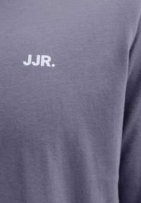 Κοντινό πλάνο μοβ υφάσματος με λευκό κείμενο "JJR." τυπωμένο στο πάνω μέρος του στήθους ενός ρούχου.