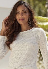 Weißer gestrickter Pullover mit Rautenmuster, rundem Ausschnitt und langen Ärmeln. Große goldene Creolen ergänzen den Look.