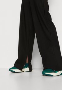 Person wearing loose black pants with side slits paired with green and white sporty sneakers standing on a white floor.