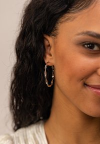 Silberne Creolen mit einem gedrehten Design, featuring eine glatte Textur und einen glänzenden Finish, getragen von einer Person mit lockigem Haar.