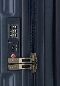 Valigia rigida blu navy con combinazione di chiusura, chiusura a cerniera e accessori metallici argentati con il nome del marchio "GUESS". Dettaglio della superficie testurizzata.