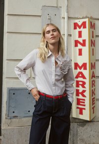 Camicia grigio chiaro con bottoni e accento floreale, abbinata a pantaloni blu scuro a vita alta e cintura rossa. Sullo sfondo si vede un'insegna.