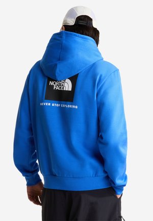 Person iført en blå hættetrøje med "The North Face"-logo og teksten "Never Stop Exploring" på ryggen, kombineret med sorte bukser og hvid kasket.
