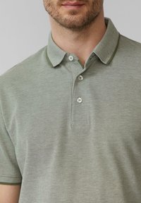 Polohemd aus gedämpftem grünem Stoff mit einem strukturierten Muster, ausgestattet mit einem einfarbigen grünen Kragen und einer Knopfleiste mit zwei Knöpfen. Kurze Ärmel.