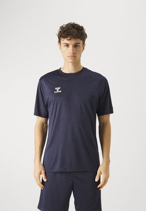 Młody mężczyzna z kręconymi włosami, ubrany w granatową sportową koszulkę Hummel i pasujące spodenki na jednolitym jasnym tle.