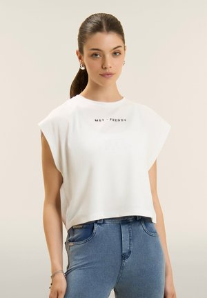 Nuori nainen, jolla on tummat taakse sidotut hiukset, pukeutuneena valkoiseen hihattomaan toppiin, jossa lukee "MST - FREDDY", ja korkeavyötäröisiin sinisiin farkkuihin.