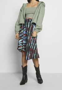 Sudadera de cremallera verde claro, top corto gris ajustado, falda de teñido anudado colorida con patrones rojos, azules y negros, y botas negras hasta el tobillo.