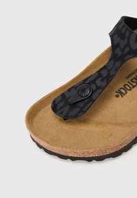 Μαύρο σανδάλι τύπου φλιπ-φλοπ με υφασμένη λουρίδα, που παρουσιάζει βάση από φελλό, μαύρη εξωτερική σόλα και κυκλικό λογότυπο στη λουρίδα.