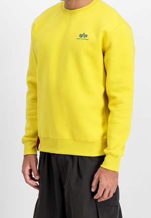 Mand iført en knaldgul Alpha Industries sweatshirt med ribstrikkede manchetter og kant, kombineret med løse sorte bukser.