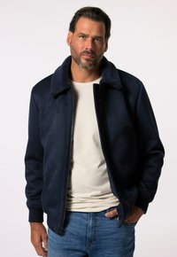 Marineblaue Kunstsuedejacke mit einem flauschigen Kragen, Reißverschluss und gerippten Bündchen, getragen über einem hellen Shirt und Jeans.