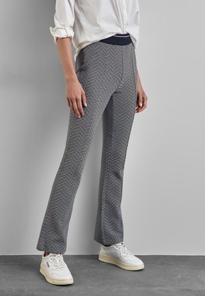 Persoon gekleed in zwart-witte gebreide broek met patroon, witte opgerolde blouse en witte sneakers, staand op een grijze vloer.