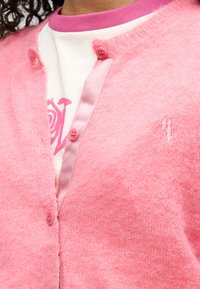 Ροζ ζακέτα με χνουδωτή υφή, που έχει κουμπιά, στρογγυλό ροζ γιακά και κεντημένη λεπτομέρεια. Φοριέται πάνω από ένα λευκό πουκάμισο με ροζ γραφικό μοτίβο.