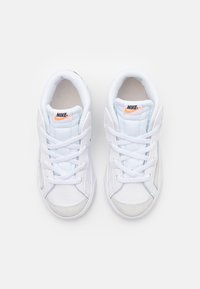 Bílé tenisky Nike s texturovanou koženou špičkou a síťovaným jazykem. Obsahují tkaničky a oranžové logo Nike na jazyku. Zaoblený design.