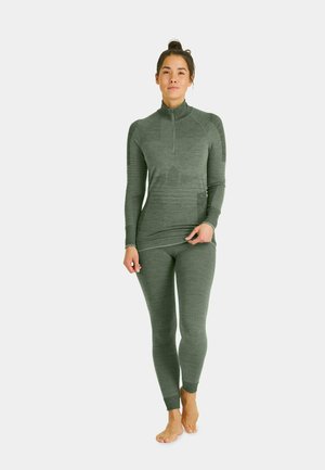 Woman standing barefoot wearing fitted green long-sleeve zip-up top and matching leggings against a plain background.