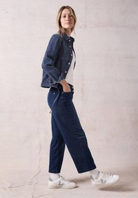 Farkkutakki tummansinisen viimeistelyn kanssa, valkoinen t-paita ja suorat tummansiniset housut. Kengät ovat valkoiset harmailla yksityiskohdilla.