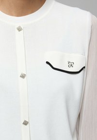 Haut en tricot blanc avec une patte de boutonnage ornée de strass carrés et une poche sur la poitrine gauche avec un contour noir et des finitions en argent.
