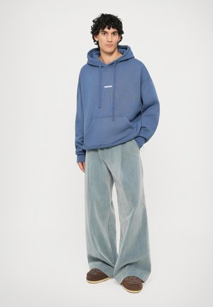 Felpa blu con cappuccio, tasca kangaroo frontale e logo bianco "BONSAI". Abbinata a pantaloni a gamba larga azzurri e scarpe marroni.