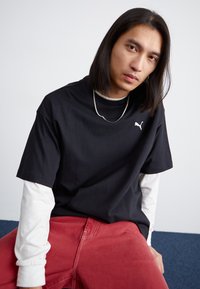 Tricou negru din bumbac cu mâneci scurte, prezentând un logo alb Puma, suprapus peste o cămașă albă cu mâneci lungi. Asortat cu pantaloni roșii.