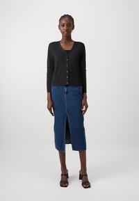 Svart strikket cardigan med V-hals og seks knapper foran, satt sammen med et denim skjørt med en frontspalte. Modellen bruker svarte hælisandaler.