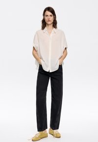 Light beige short-sleeve button-up shirt with a textured grid pattern, paired with black loose-fit trousers and olive green suede shoes.