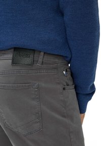 Grijze broek met een gestructureerde afwerking, voorzien van een zwart lederen patch met het label "Brax" op de tailleband. Blauwe gebreide trui gedeeltelijk zichtbaar.