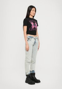Svart avkortet t-skjorte med rosa sommerfugl grafikk og tekst «VERSACE JEANS COUTURE», kombinert med lyse vasket smale jeans og svarte støvler.