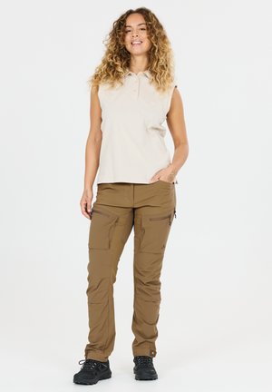 Smilende kvinde med krøllet hår iført ærmeløs beige polotrøje, brune cargobukser og sorte vandresko, stående med den ene hånd i lommen.