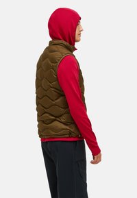 Bruine puffervest met een gewatteerd patroon, gecombineerd met een rode structuur lange mouwtop. Het vest heeft een hoge kraag en wordt gedragen over zwarte broeken.