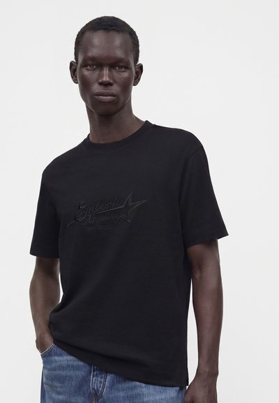 Άνδρας που φοράει μαύρο κοντομάνικο t-shirt Emporio Armani και μπλε τζιν, στέκεται μπροστά σε λευκό φόντο.