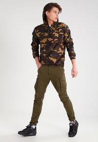 Sudadera con capucha de camuflaje con un bolsillo frontal, con tonos de verde y marrón; combinada con pantalones cargo oliva y zapatillas negras.