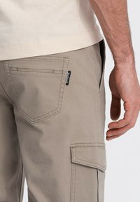 Beige cargobroek gemaakt van stevig materiaal, met patiopockets, gestikte details en een klein zwart label aan de zijkant.