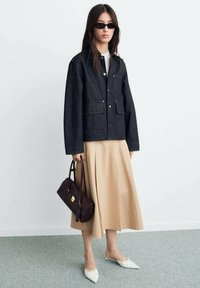 Dark denim jacket with button closure and two front pockets, paired with a beige midi skirt. White pointed flats and a dark handbag complete the look.