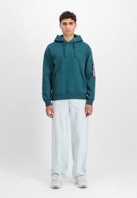 Türkise kapuutsviiter esiosaga tasku ja logopatchiga. Koos helesiniste laiemate pükstega ja hallide tossudega. Punane sildi detail varrukal.