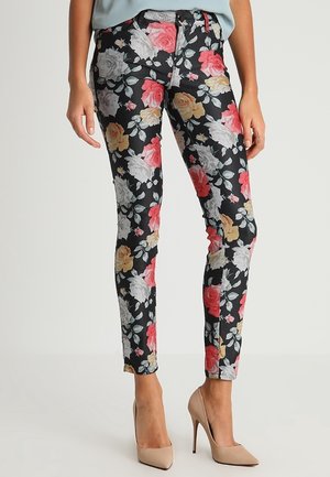 Zwarte skinny broek met rode, gele en witte rozenpatronen, gedragen met beige hoge hakken en een lichtblauwe top, staand tegen een effen achtergrond.