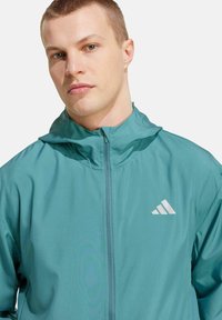 Chaqueta cortavientos en color verde azulado con capucha, confeccionada en material ligero, que cuenta con un cierre frontal y un logo reflectante en el pecho.