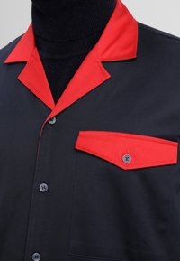 Kortärmad skjorta med svart kropp, röd krage och bröstficka, tillverkad av ett mjukt tyg med knappstängning.