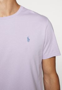 Světle fialové bavlněné tričko s krátkými rukávy a kulatým výstřihem, které má na levé části hrudi malé modré vyšívané logo.