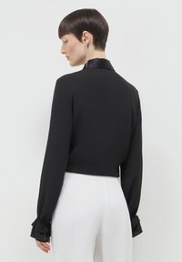 Blouse noire avec un col en satin, manches longues avec des poignets à volants, et un design court. Portée avec un pantalon blanc taille haute.