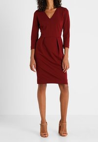 Bordowa sukienka bodycon z dekoltem w serek, trzyczwartowymi rękawami i plisowaniem w talii. Noszona z brązowymi sandałami na wysokim obcasie.