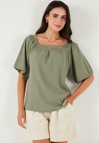 Sage green blouse with a square neckline, intricate lace detailing, puffed sleeves, and a lightweight texture, paired with beige shorts.