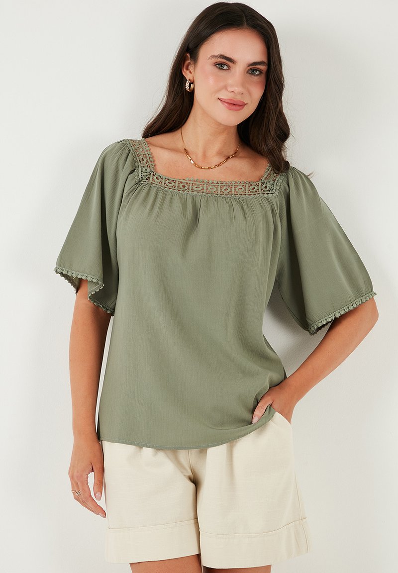 Sage green blouse with a square neckline, intricate lace detailing, puffed sleeves, and a lightweight texture, paired with beige shorts.