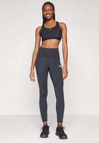 Χαμογελαστή γυναίκα με πλεξούδες, φορώντας μαύρο αθλητικό σουτιέν, σκούρες γκρι κολάν και μαύρα αθλητικά παπούτσια, στέκεται μπροστά σε απλό φωτεινό φόντο.