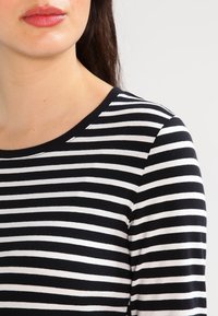 Kvinna som bär en svartvit randig långärmad skjorta, visar sin hals, läppar och övre axel mot en enkel bakgrund.