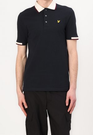 Granatowa koszulka polo z kontrastującym kremowo-bordowym kołnierzykiem, plisa na trzy guziki oraz małym żółtym logo na lewej piersi. Krótkie rękawy.