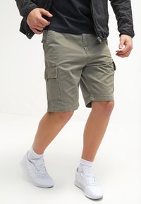 Pantalones cortos de cargo color caqui, hechos de tejido de algodón, con múltiples bolsillos, un corte relajado y un diseño de pierna recta. Combinados con zapatillas blancas.