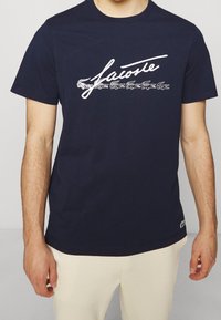 Navy cotton t-shirt featuring white Lacoste logo with crocodile graphic. Short sleeves and standard fit, smooth texture, logo on hem.