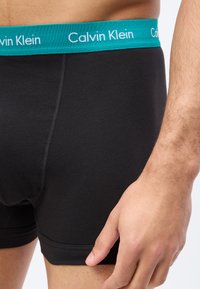 Férfi fekete boxeralsót visel világoskék Calvin Klein derékpánttal, jobb keze lazán oldalt, egyszínű háttér előtt.