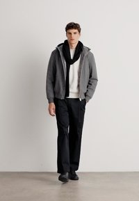 Siva zip-up jakna s kapuco, bela teksturirana pulover, črne hlače in črn šal, ki jih nosi moški model proti nevtralni ozadju.