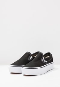 Zapatillas de lona negras sin cordones con suelas de goma blanca y detalles de costura. Presentan una punta redonda y paneles elásticos en los lados para mayor comodidad al ponérselas.